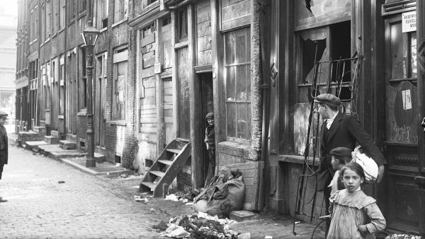 Een zwart-witfoto van een arbeiderswijk begin 20ste eeuw in Amsterdam, met vervallen woningen, troep op straat en een aantal mensen die op straat lopen