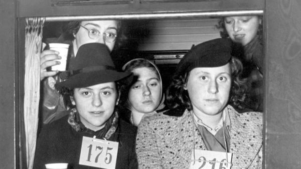 Een zwart-witfoto van Joodse vluchtelingen die uit het raam van een trein kijken met bekertjes in hun hand en registratienummers op hun borst.