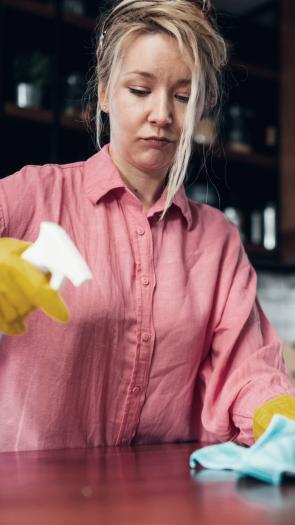 Een vrouw kijkt naar het aanrecht terwijl ze aan het schoonmaken is met handschoenen aan, een doekje in haar hand en een spray in de andere hand