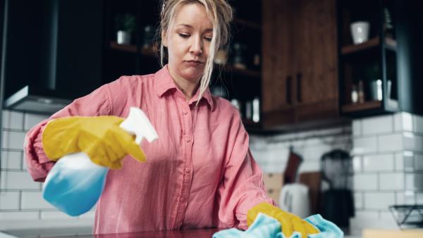 Een vrouw kijkt naar het aanrecht terwijl ze aan het schoonmaken is met handschoenen aan, een doekje in haar hand en een spray in de andere hand