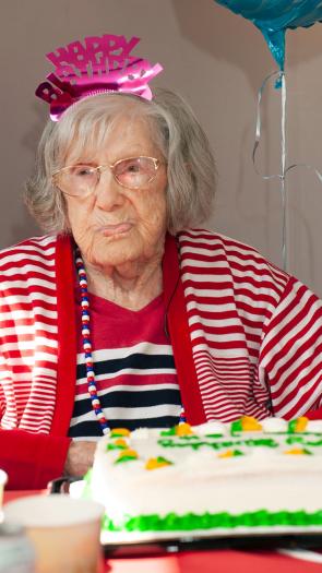 Een oude vrouw staart voor zich uit met een 'happy birthday' dingetje op haar hoofd en een taart voor zich