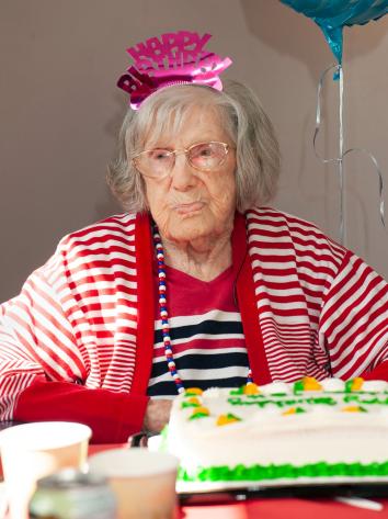 Een oude vrouw staart voor zich uit met een 'happy birthday' dingetje op haar hoofd en een taart voor zich
