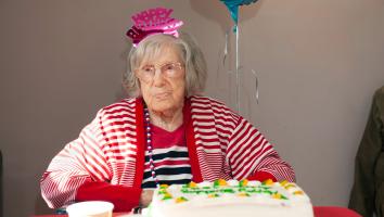 Een oude vrouw staart voor zich uit met een 'happy birthday' dingetje op haar hoofd en een taart voor zich