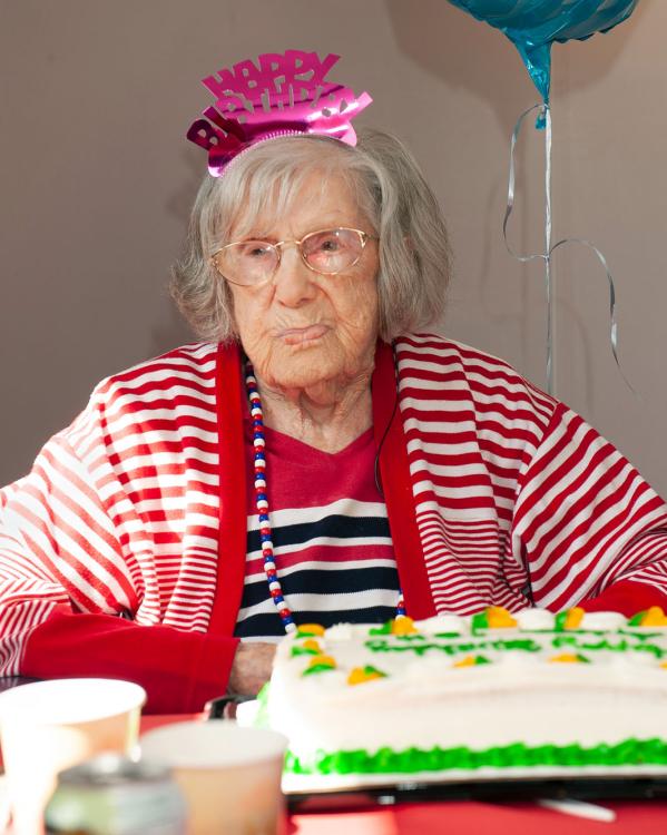 Een oude vrouw staart voor zich uit met een 'happy birthday' dingetje op haar hoofd en een taart voor zich