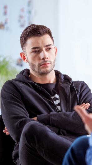 Een man zit met zijn armen over elkaar voor zich uit te staren terwijl een man die met de rug naar de camera zit iets vertelt