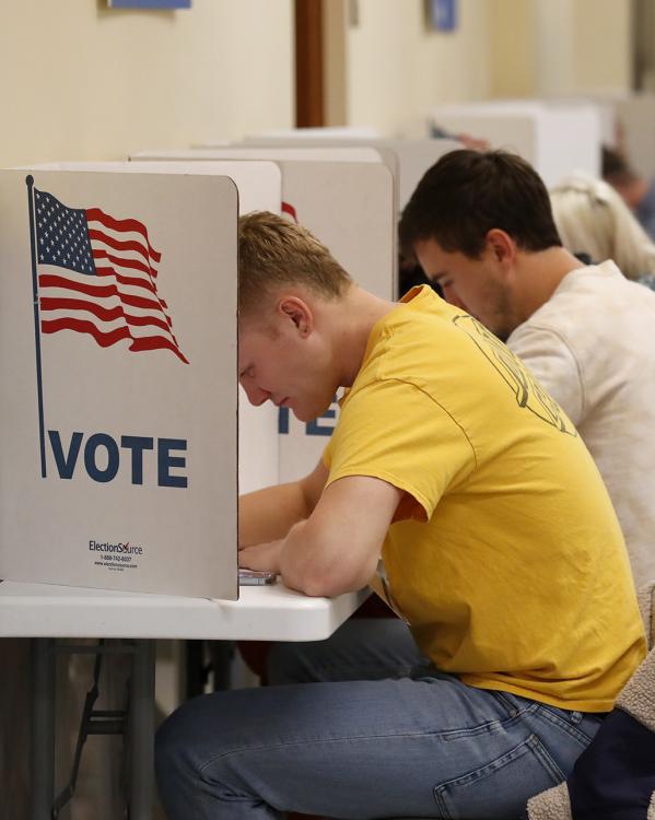 Rij jongeren zit aan tafels met afgeschermde zijkanten waar de Amerikaanse vlag op staat met de tekst 'Vote'. Ze schijven.