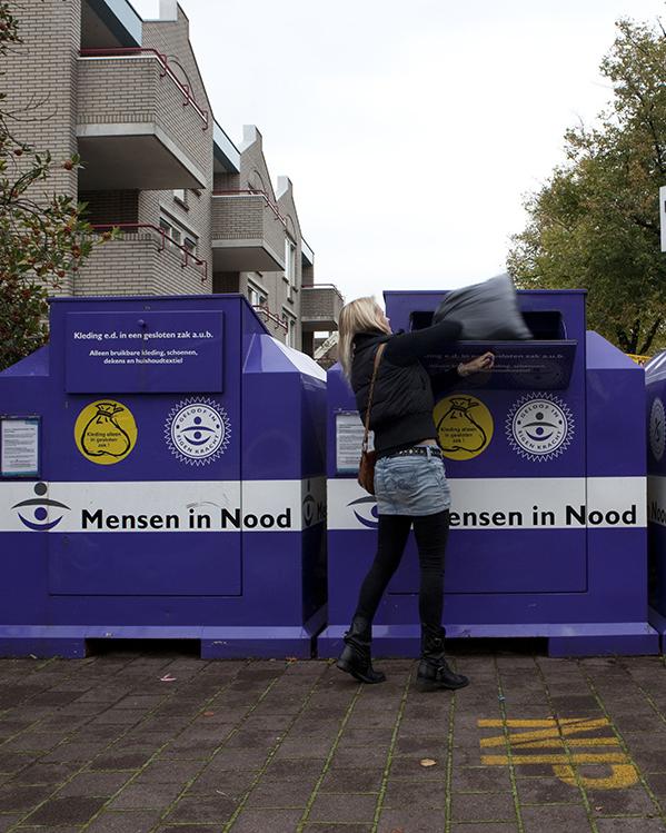 Vrouw gooit vuilniszak ik een container met de tekst 'Mensen in nood' erop gedrukt. De container is onderdeel van een rij containers bedoeld voor kleding