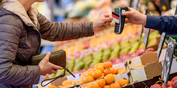 Iemand rekent af met de pinpas bij een mobiel pinautomaat op de markt, met op de achtergrond de marktkraam van de groenteboer