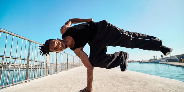 Een breakdancer doet een pose waarbij hij liggend in de lucht hangt met één hand op de grond. Hij doet dit op de stoep, verder zie je een blauwe lucht en water op de achtergrond.