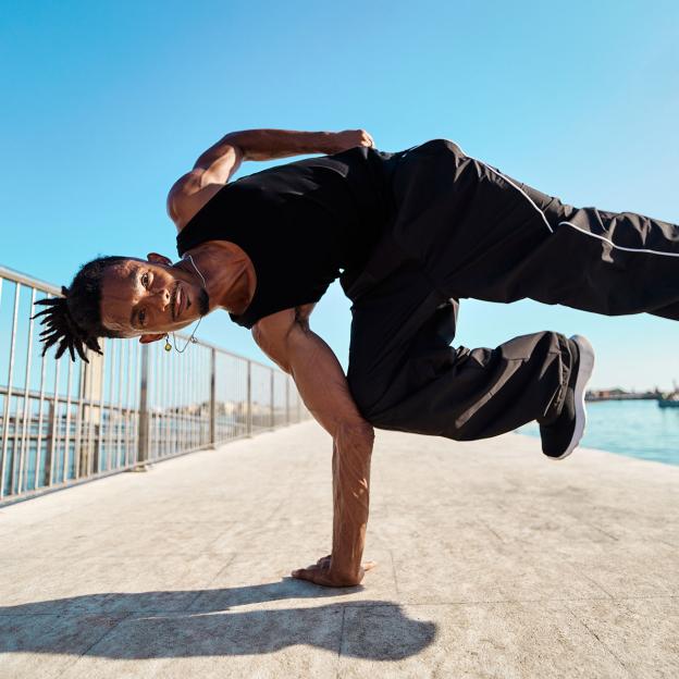Een breakdancer doet een pose waarbij hij liggend in de lucht hangt met één hand op de grond. Hij doet dit op de stoep, verder zie je een blauwe lucht en water op de achtergrond.